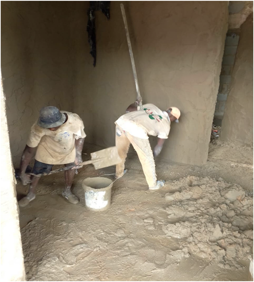 A photograph of two workers plastering an interior wall with wet cement using hand tools, without wearing protective gear.