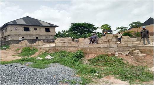 A photograph of workers laying concrete blocks on a building site without PPE, surrounded by unfinished walls and greenery.