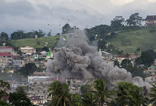 A large explosion produces a dense cloud of smoke rising above buildings. Debris is visible in the air. Nearby structures are partially obscured.
