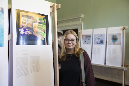 A young woman stands beside an exhibition display board presenting artwork and written content during The Belonging Project event.