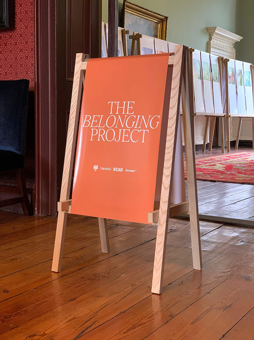 An exhibition sign for The Belonging Project stands on a wooden floor beside display panels in a gallery interior.