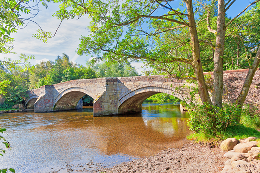 A stone arch bridge with three spans over a shallow river surrounded by trees.