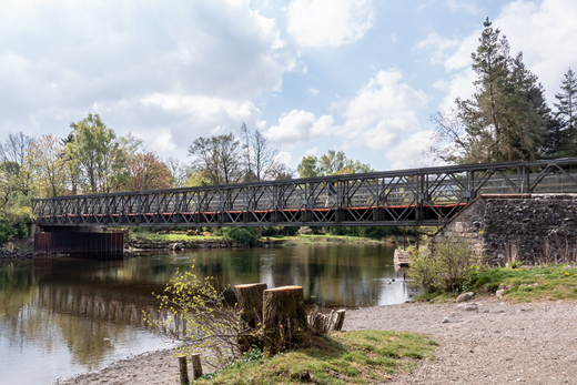 A metal truss bridge crossing a river with trees and a gravel bank nearby.