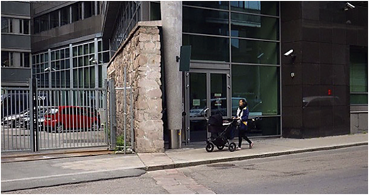 A photograph shows a woman pushing a stroller past a modern glass building and a fenced parking area.