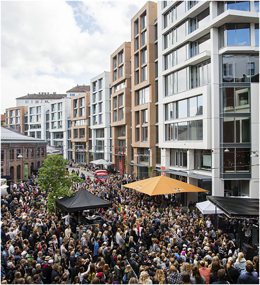 A photograph shows a large crowd filling a street lined with tall modern buildings and outdoor canopies.