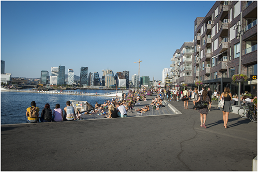A photograph shows crowds sitting and walking along a waterfront promenade beside modern high-rise buildings.