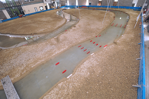 A large model of a riverbed is shown, featuring gravel, sand, and red markers on a smooth surface representing water flow.
