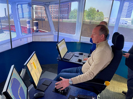 A person operates a boat simulator in a control room with multiple screens displaying navigation data, while another individual observes in the background.