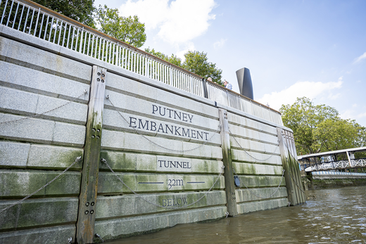A river embankment wall with inscriptions of Putney Embankment, Tunnel, and 32 m Below, surrounded by trees and a boat nearby.