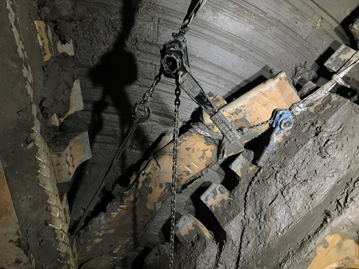 The image shows a large, mechanical structure with chains and equipment inside a dark, possibly underground environment, surrounded by wet concrete and dirt.
