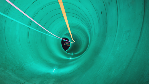 A smooth circular tunnel extends into the distance. Long exposure lights form curved lines running through the centre toward a dark opening. The tunnel's inner surface is even and continuous, showing faint joint lines between segments. The view looks straight through the tunnel, creating a sense of depth.