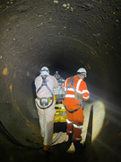 Workers inspect the interior of a large underground tunnel using portable equipment and lighting.