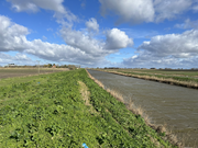 Landscape of a river or drainage channel beside grassy embankments under a cloudy sky.