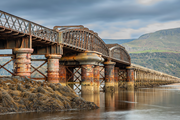A railway truss bridge extends across water on cylindrical piers with multiple spans aligned toward distant hills.