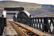 A railway track runs through a steel truss bridge that spans above water with repeating arch frames aligned toward distant hills.