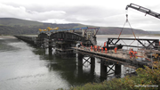 A bridge construction above water shows workers on a deck platform while a crane lifts a truss component near a span supported by scaffolding.