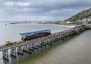 A railway bridge above water shows workers along the deck and a transported truss span positioned on the bridge structure.