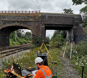 A person wearing safety gear operates a laptop near a railway bridge with cameras and data equipment.
