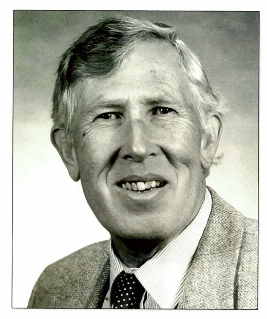 A black-and-white portrait of a smiling man with grey hair, wearing a suit jacket over a collared shirt with a tie.