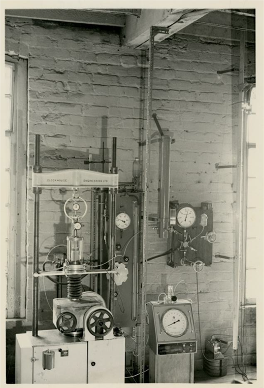 A black and white photo of a laboratory setup featuring various scientific instruments, including pressure gauges and a mechanical apparatus, set against a brick wall.