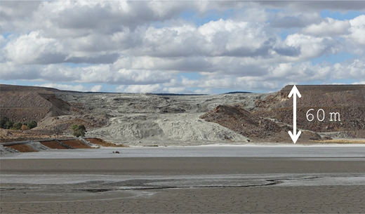 A large, barren landscape features a steep, grey hill rising approximately sixty metres. The sky is partly cloudy above the scene.