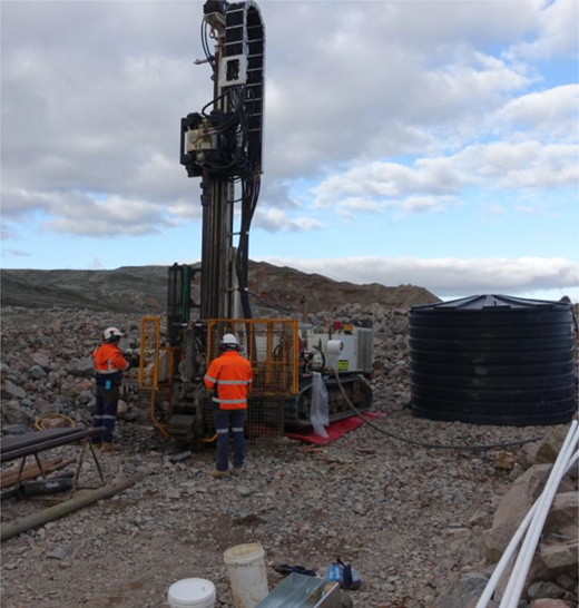A drilling rig stands amidst rocky terrain, with workers in safety gear nearby, showcasing a mining operation site.
