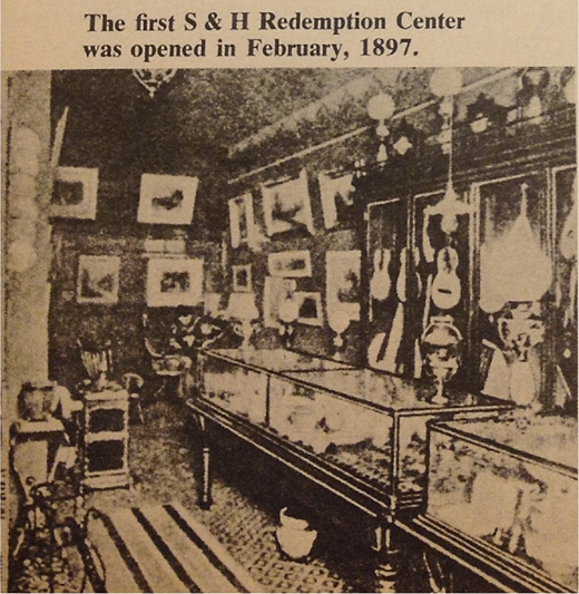 A view of the first S and H redemption centre opened in February 1897, showing interior display cases, framed pictures, hanging instruments, and household items arranged along walls.