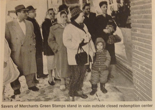 A group of adults and a child stand outside a brick building. The people wear winter coats, hats, and scarves. Snow is visible on the ground.