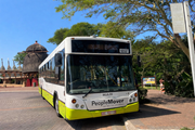 A People Mover bus with 10537 displayed is parked beside a paved walkway surrounded by trees and buildings in an outdoor urban setting.