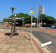 A bus stop shelter and roadside sign stand beside a curved roadway near a roundabout and trees with an arrow pointing to the pavement.