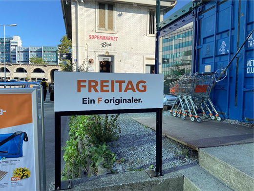 A rectangular outdoor sign displaying the adapted logo and slogan FREITAG Ein F originaler, mounted on a frame near a supermarket entrance with shopping trolleys and surrounding buildings visible.