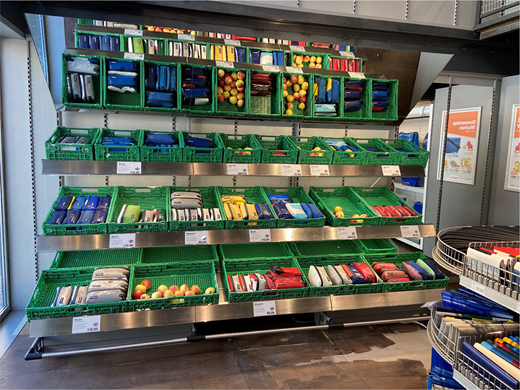 A store display in a FREITAG shop arranged like supermarket shelving, with crates holding wallets, pouches and bags presented like groceries, referencing Migros.