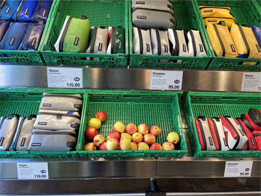 Supermarket-style shelving with plastic crates displaying FREITAG pouches and bags labelled Wayne at 110.00 and Cheyenne at 95.00, alongside loose apples arranged in a crate.