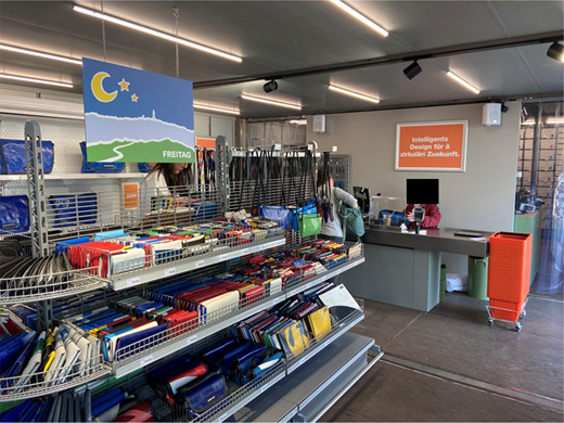 Interior of a FREITAG store with supermarket-style metal shelving displaying wallets and bags, a hanging landscape sign with crescent moon and stars labelled FREITAG, a checkout counter with computer and wall sign reading Intelligents Design für a zirkuläre Zukunft, hanging straps, overhead strip lighting, and a shopping trolley beside the till.