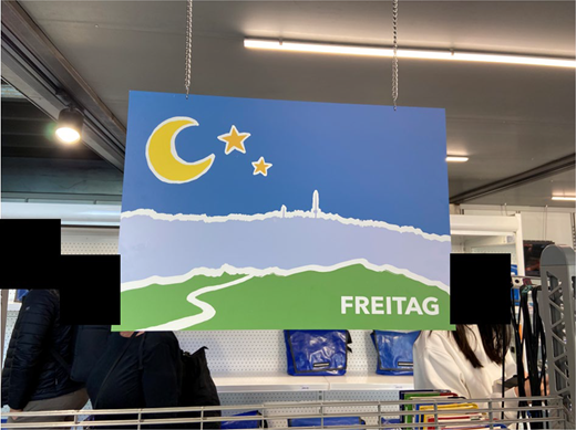 A hanging rectangular sign inside a FREITAG store showing a crescent moon, two stars, a landscape with a winding path and the word FREITAG.