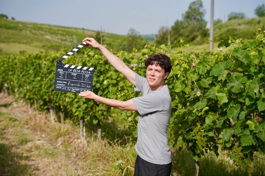 A person stands among vineyard rows while holding a clapperboard for La boa rosa, roll 2, scene 2, take 2.