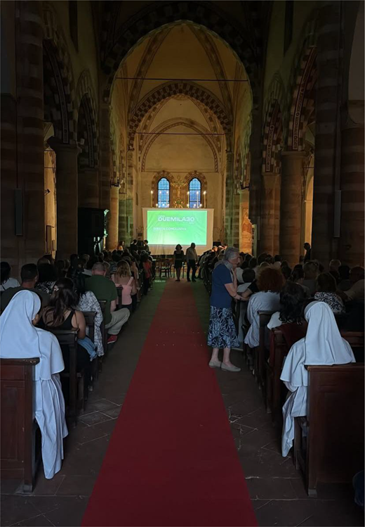 A gathering in a church features a projection screen displaying text, with an audience seated on wooden benches along a carpet.