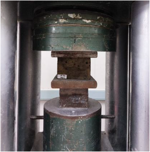 A photo shows a compression testing machine applying vertical force to a rectangular concrete block sample.