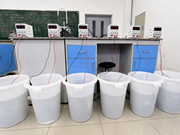 A laboratory setup shows six D C power supplies connected by cables to six plastic buckets.