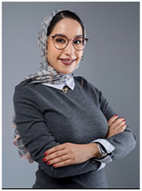 A woman with glasses and a headscarf standing with arms crossed.