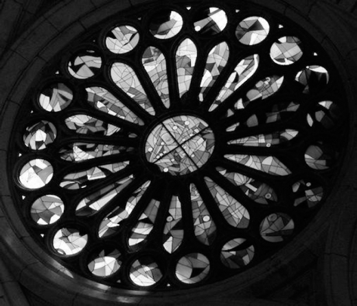 A circular window with a central pane surrounded by petal like segments arranged in radial symmetry. Each segment contains geometric stained glass patterns framed by stone tracery, forming a detailed Gothic-style architectural design.