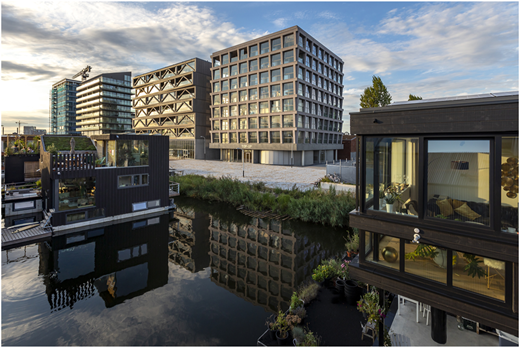 A photograph shows modern waterfront buildings reflected in a calm canal under a partly cloudy sky.