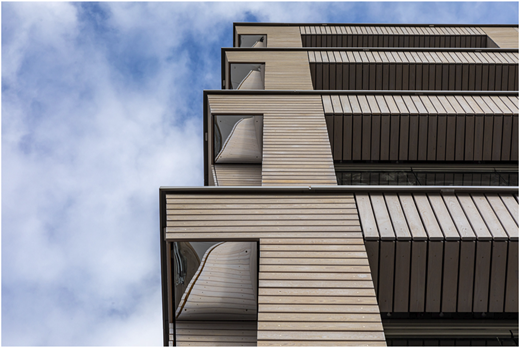 A photograph shows an upward view of a timber-clad building with stacked projecting balconies against a cloudy sky.
