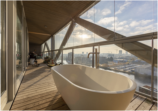 A photograph shows a freestanding bathtub on a timber balcony enclosed with glass, overlooking a cityscape.