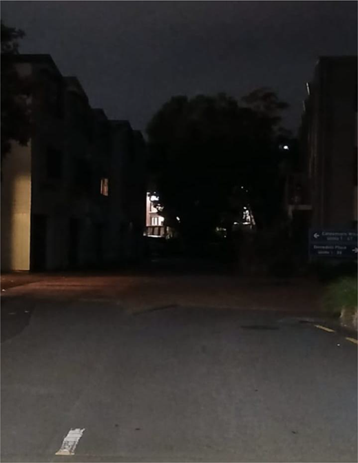 A dimly lit road curves between buildings and dense trees, with an illuminated entrance and a directional sign beside the junction.