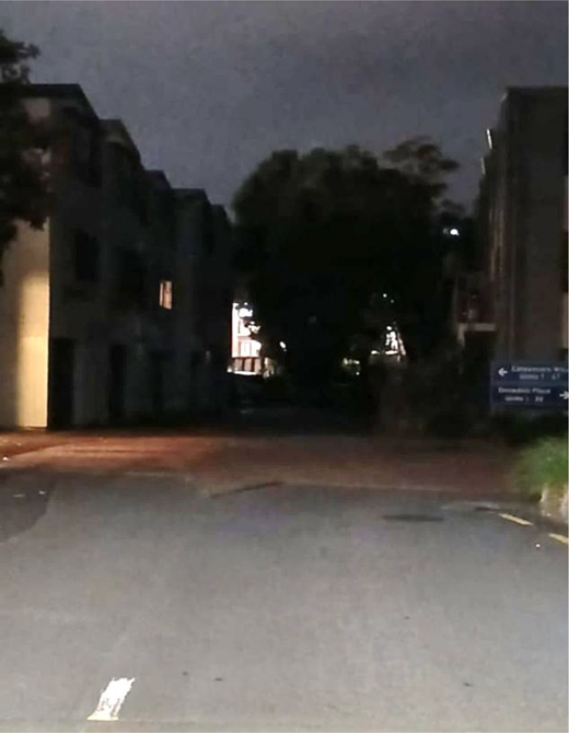 A dim road curves between buildings and trees, with an illuminated entrance, distant lights, a directional sign and a short foreground road marking.