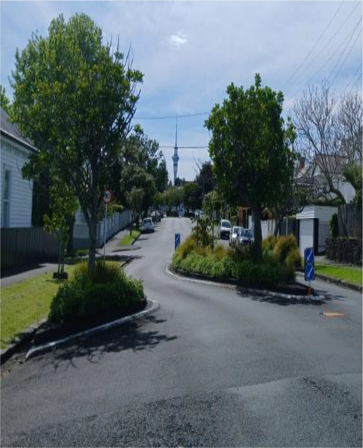 A residential street curves around planted traffic islands, with houses, parked vehicles, trees, roadside signs and a distant tower.