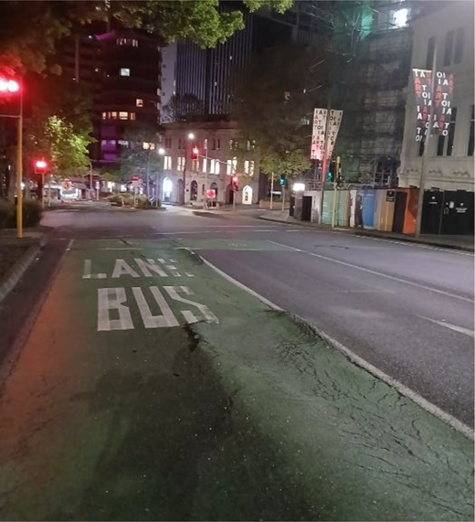 A night-time bus lane leads towards an intersection with traffic lights, buildings, trees, roadworks and illuminated windows.