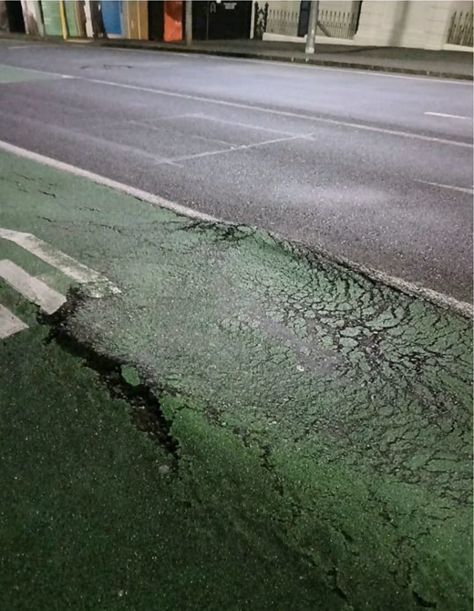 A cycle lane beside a road has extensive cracking, broken surface edges and worn lane markings.