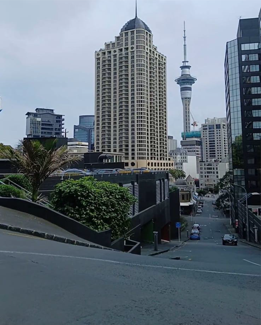 A steep city street descends between high-rise buildings, with vehicles, planted terraces and a tall observation tower in the distance.